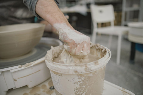 a hand holding a fistful of wet clay above a white bucket with clay residue on the rim and sides